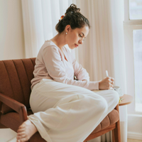 A woman in lounge pants sitting on a chair and writing in a journal